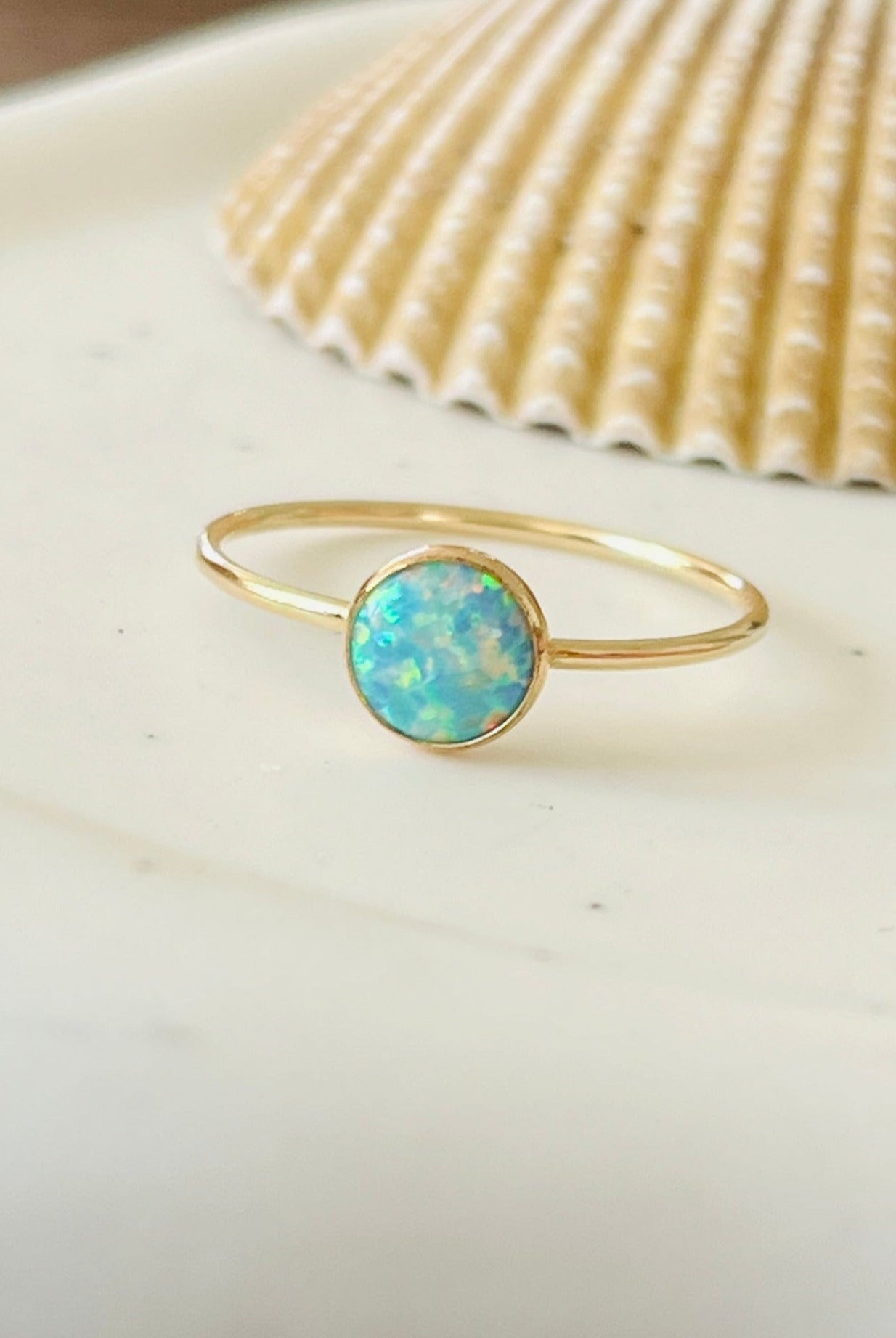 A gold ring with a seafoam opal center stone, placed on a white dish with a seashell and pink stone in the background.