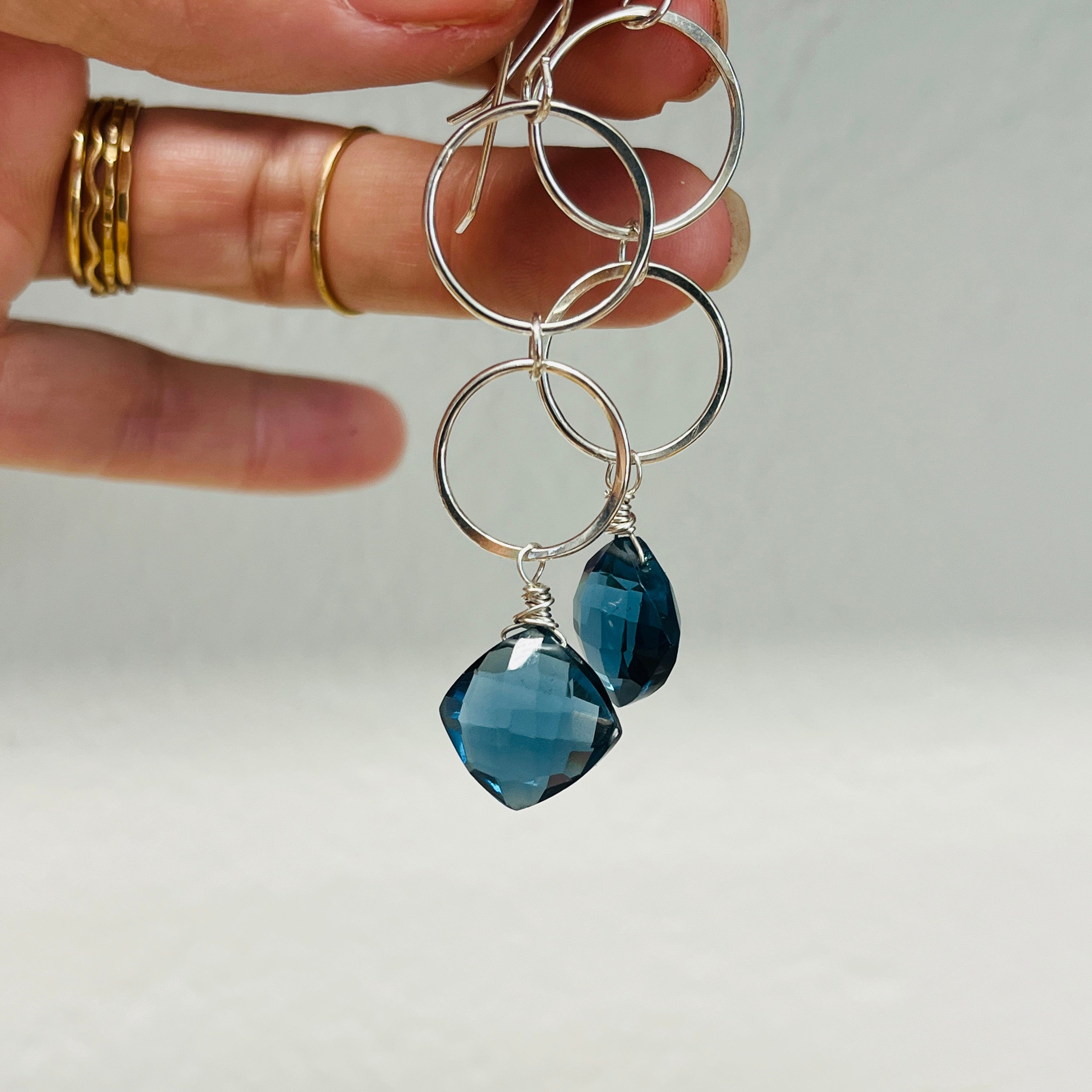 Silver earrings with blue heart-shaped beads held by a hand against a neutral background