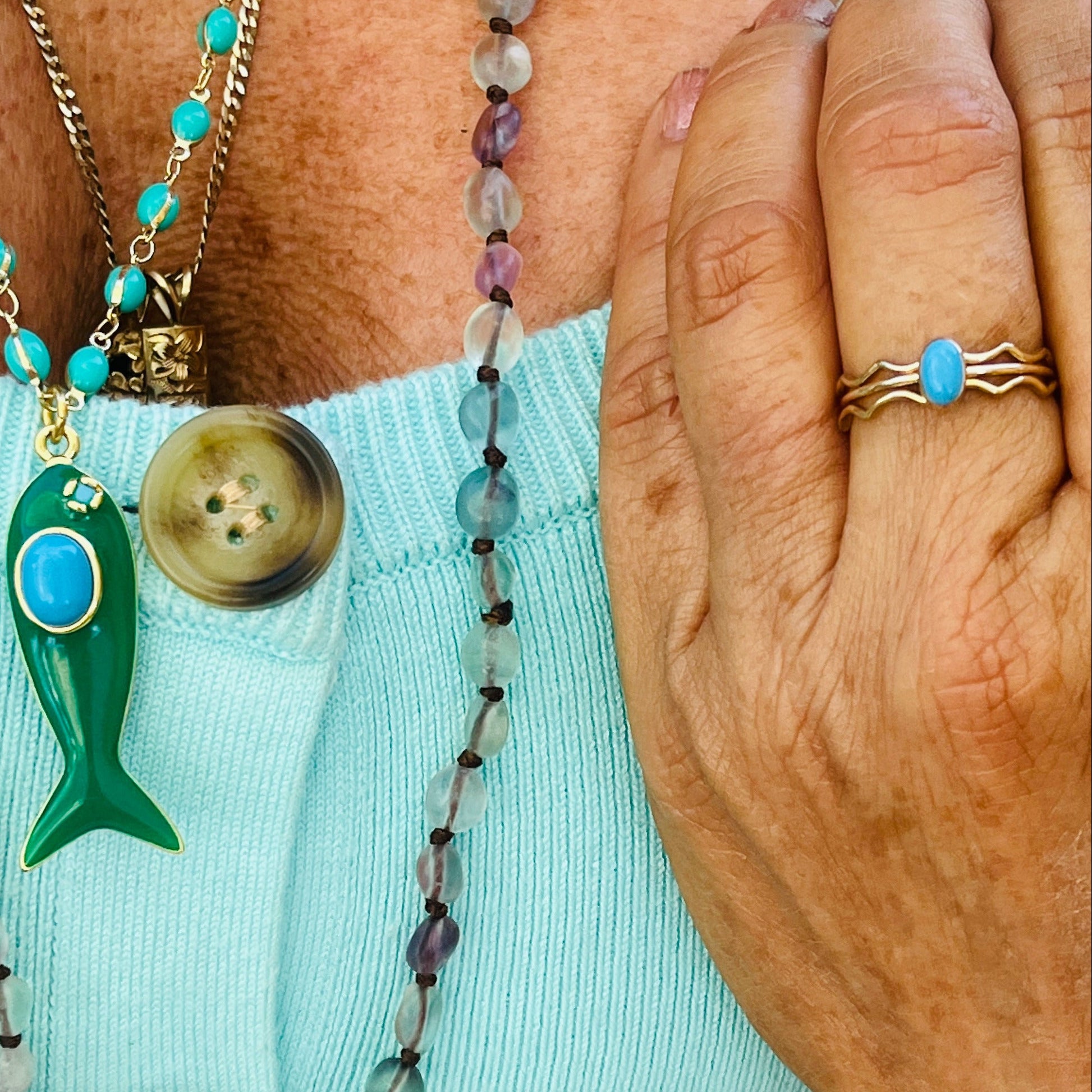 Close-up of a person wearing multiple necklaces with beads and buttons, and a ring on a light blue background.