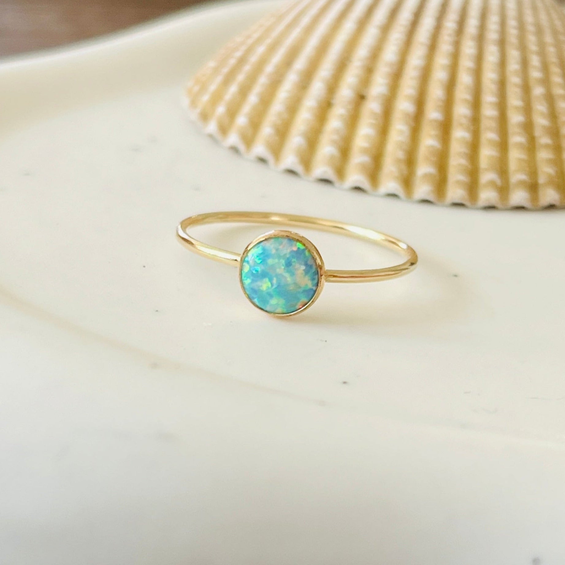 A gold ring with a seafoam opal center stone, placed on a white dish with a seashell and pink stone in the background.